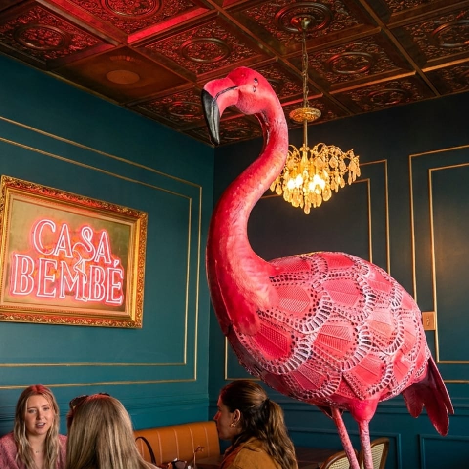 A large pink flamingo statue stands in a restaurant with teal walls, a neon sign, and patrons.
