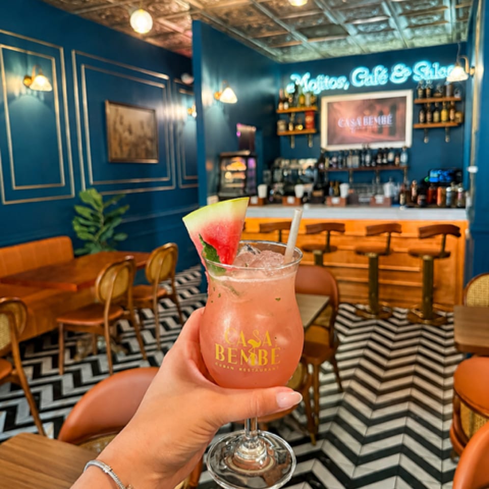 Hand holding a pink Casa Bembe cocktail with watermelon in a vibrant blue restaurant interior.